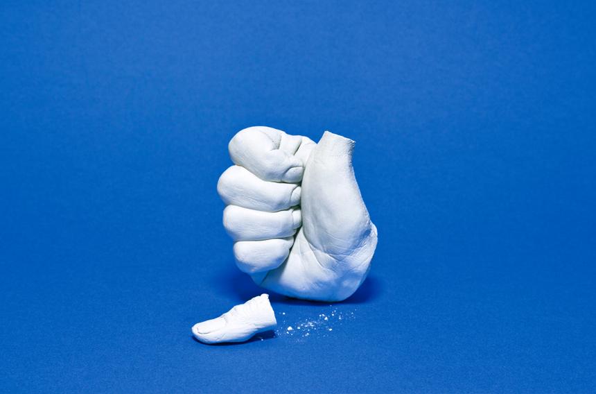 Une oeuvre de Catherine Radosa intitulée "Pollice Incerto". 2015. Une main grise sculptée, en plâtre, sur fond bleu. Elle fait le geste du pouce en l'air. Le pouce de la main est brisé et repose à côté d'elle.